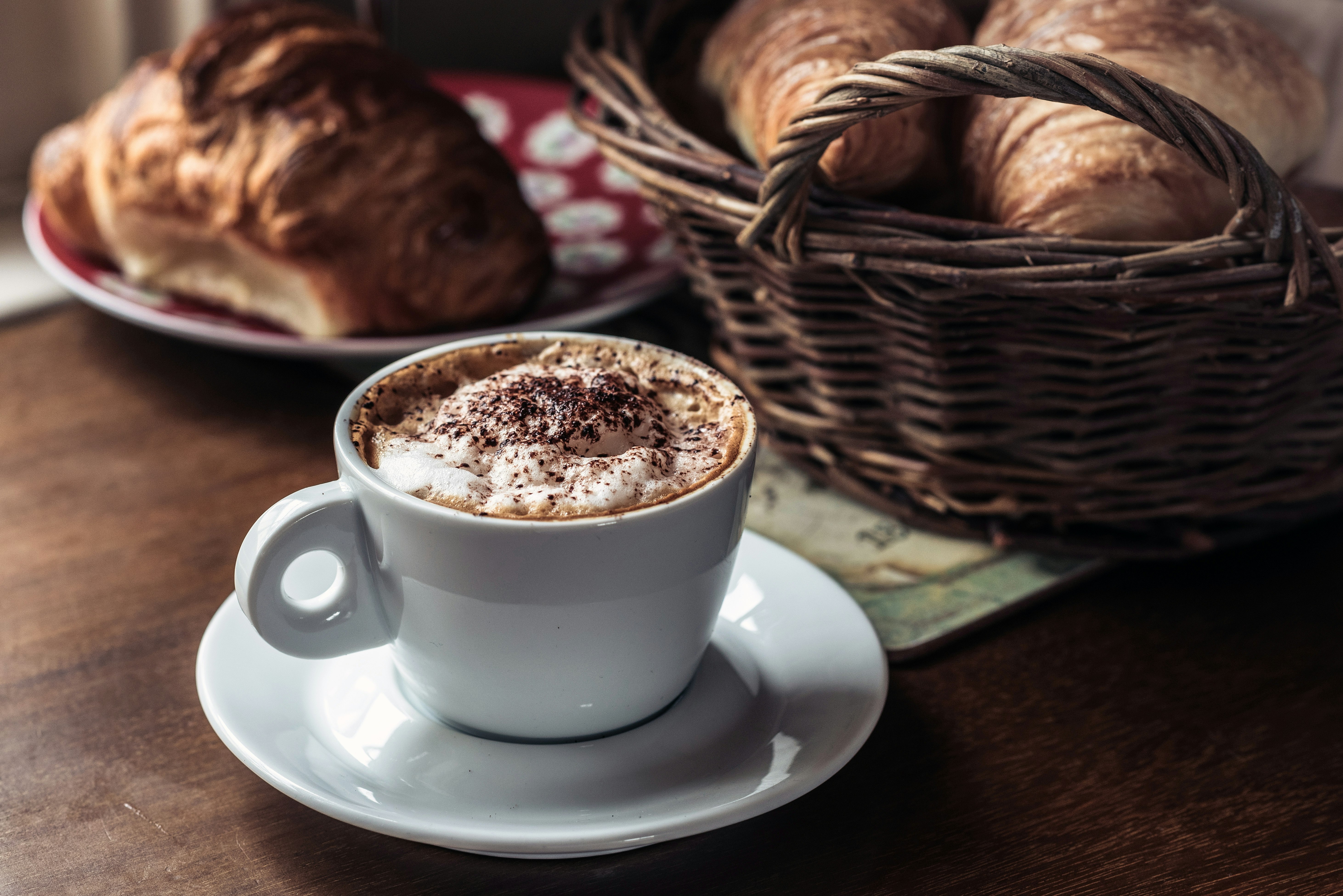 capuccino coffee on white ceramic cup with saucer beside pastry bread in basket and red plate